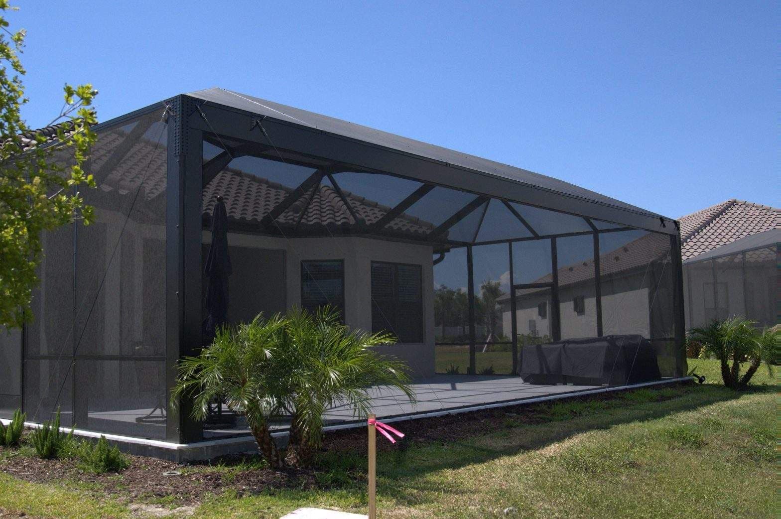 A spacious screened patio enclosure beside a home, with a covered roof, outdoor furniture, palm plants, and bright sunny skies.