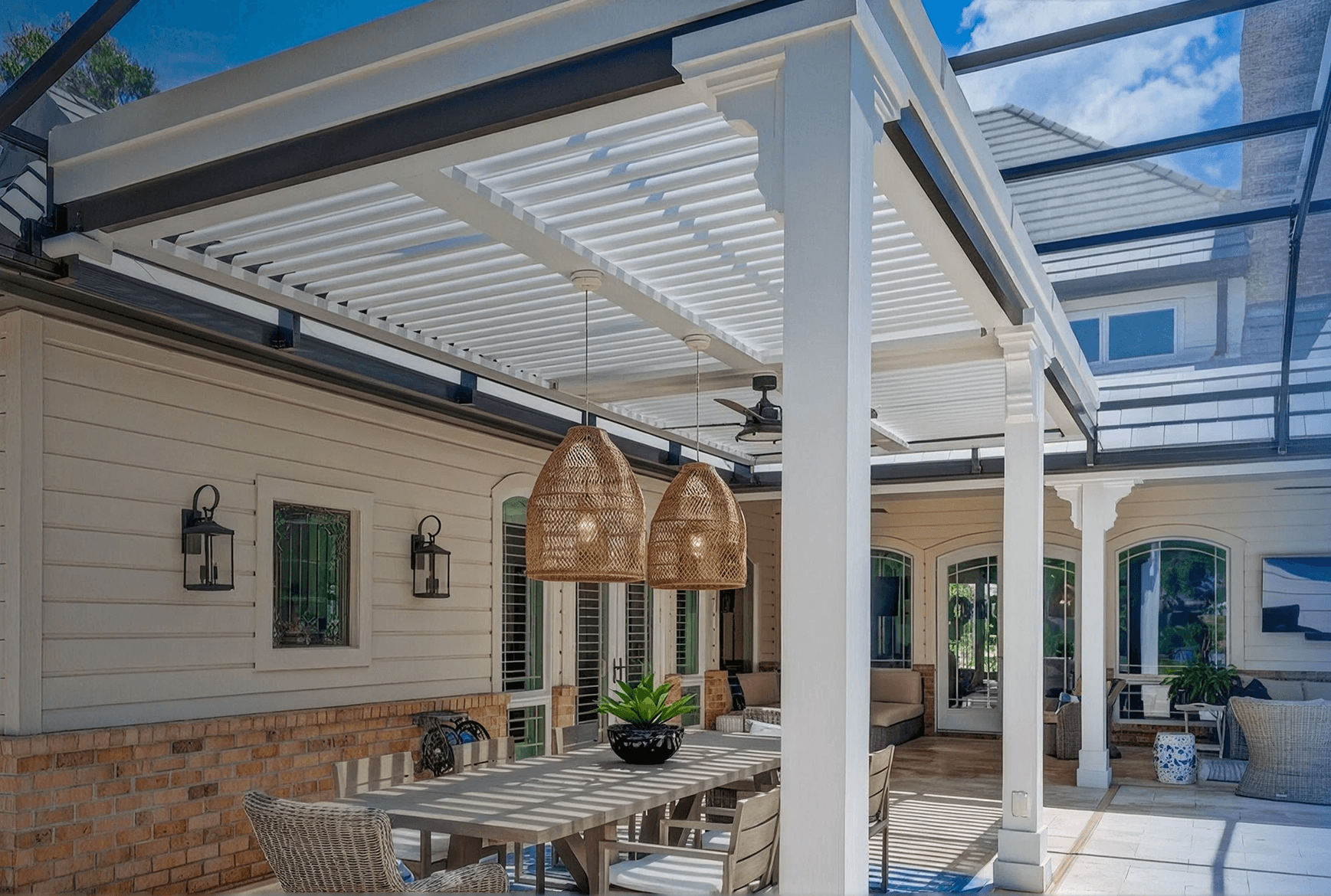 Modern patio with a slatted pergola, wicker furniture, and pendant lights. Bright, airy space with a dining table and lush plants, exuding relaxation.