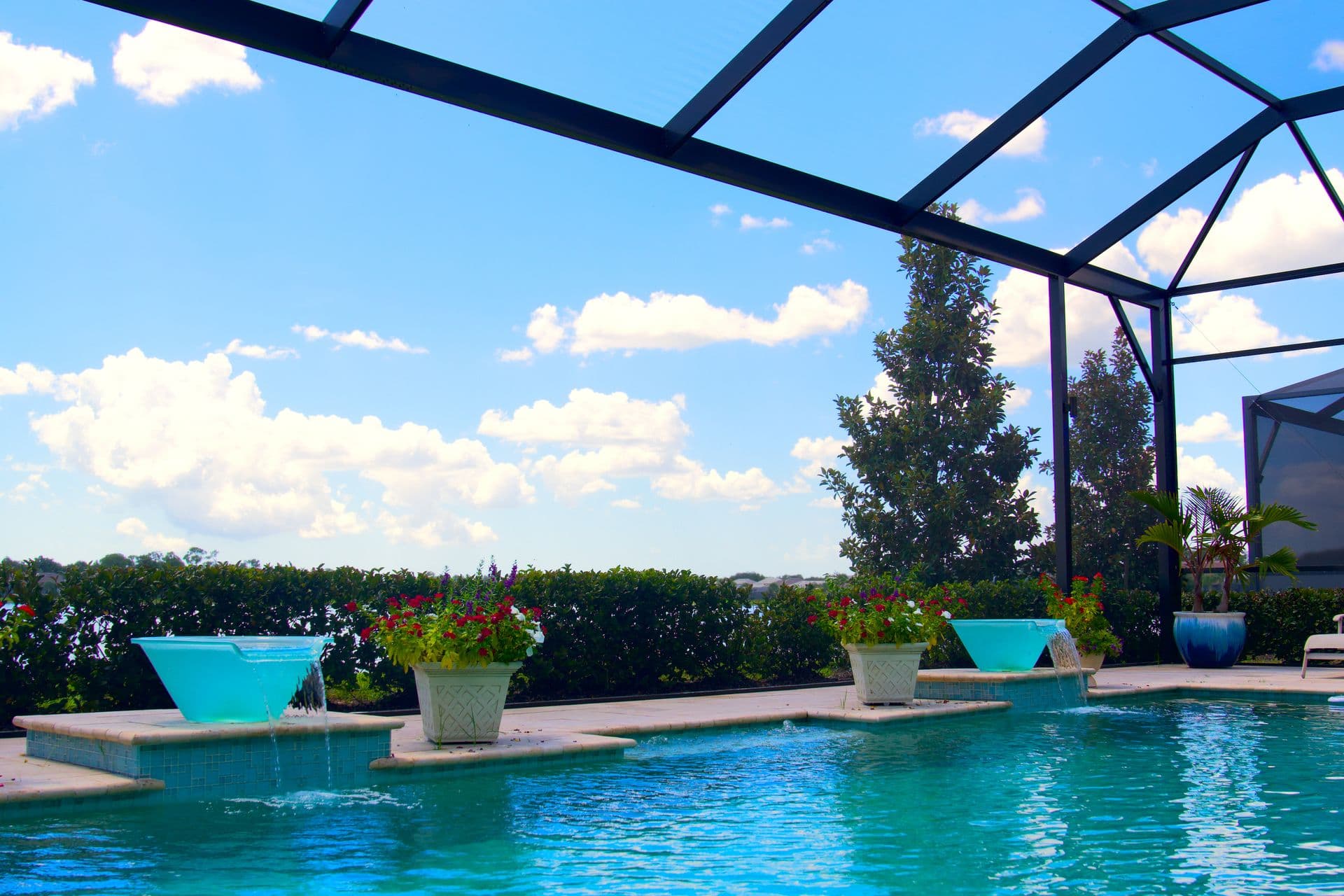 Covered patio with modern seating, a fire pit, and potted plants. Overhead slatted pergola provides shade. Greenery and open sky create a serene atmosphere.